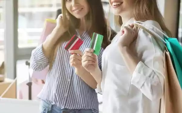 Two women holding shopping bags in one hand and a credit card in the other.