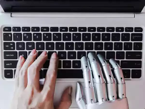 Human left hand and robotic right hand typing on a laptop computer.