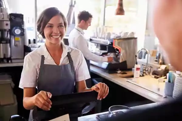 Person ordering an item from a coffee shop from a woman in a blue apron smiling.