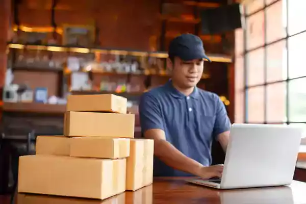 Man typing on a laptop with a pile of packages next to him.